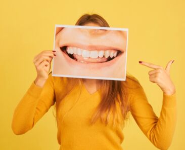 young woman holding pointing finger at picture of mouth smiling showing her teeth on yellow background. Dentist concept